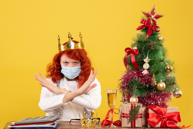 Ärztin der Vorderansicht, die mit Weihnachtsgeschenken sitzt und Krone auf gelbem Hintergrund trägt