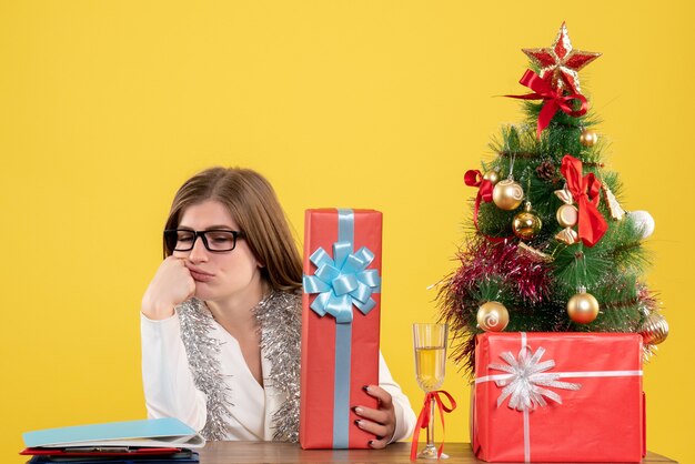 Ärztin der Vorderansicht, die mit Weihnachtsgeschenken und Baum auf gelbem Hintergrund sitzt