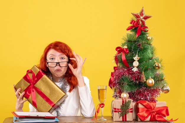 Ärztin der Vorderansicht, die mit Weihnachtsgeschenken und Baum auf gelbem Schreibtisch sitzt