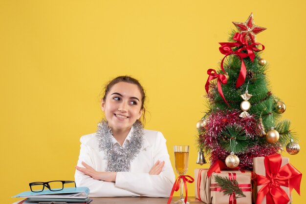 Ärztin der Vorderansicht, die um Weihnachtsgeschenke und Baum auf gelbem Hintergrund sitzt