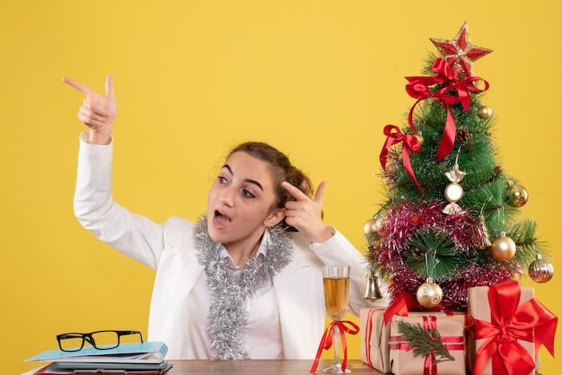 Ärztin der Vorderansicht, die um Weihnachtsgeschenke und Baum auf gelbem Hintergrund sitzt