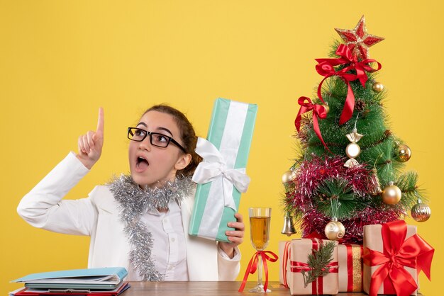 Ärztin der Vorderansicht, die um Weihnachtsgeschenke und Baum auf gelbem Hintergrund sitzt