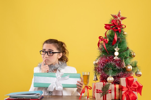 Ärztin der Vorderansicht, die um Weihnachtsgeschenke und Baum hält Geschenk auf gelbem Hintergrund sitzt