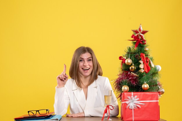 Ärztin der Vorderansicht, die vor ihrem Tisch auf gelbem Hintergrund mit Weihnachtsbaum und Geschenkboxen sitzt