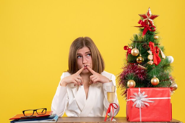 Ärztin der Vorderansicht, die vor ihrem Tisch auf gelbem Hintergrund mit Weihnachtsbaum und Geschenkboxen sitzt