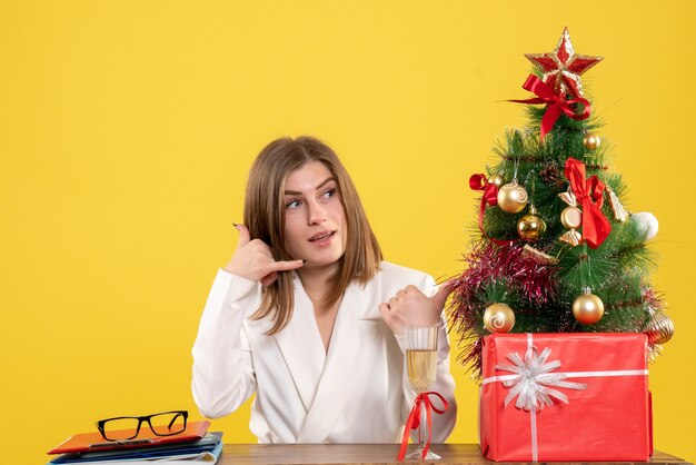 Ärztin der Vorderansicht, die vor ihrem Tisch auf gelbem Hintergrund mit Weihnachtsbaum und Geschenkboxen sitzt