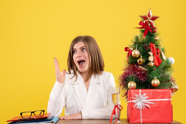 Ärztin der Vorderansicht, die vor ihrem Tisch auf gelbem Hintergrund mit Weihnachtsbaum und Geschenkboxen sitzt