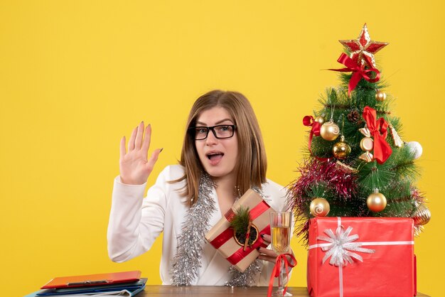 Ärztin der Vorderansicht, die vor ihrem Tisch auf gelbem Hintergrund mit Weihnachtsbaum und Geschenkboxen sitzt