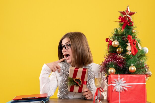 Ärztin der Vorderansicht, die vor ihrem Tisch hält Geschenk auf gelbem Hintergrund mit Weihnachtsbaum und Geschenkboxen hält