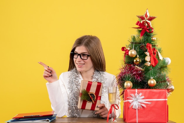 Ärztin der Vorderansicht, die vor ihrem Tisch hält Geschenk auf gelbem Schreibtisch mit Weihnachtsbaum und Geschenkboxen hält