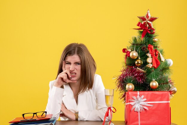 Ärztin der Vorderansicht, die vor ihrem Tisch sitzt, betont auf gelbem Hintergrund mit Weihnachtsbaum und Geschenkboxen