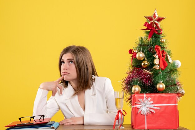 Ärztin der Vorderansicht, die vor ihrem Tisch sitzt und auf gelbem Hintergrund mit Weihnachtsbaum und Geschenkboxen denkt