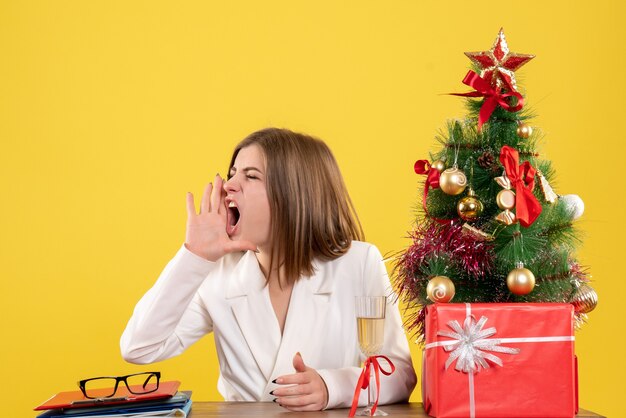 Ärztin der Vorderansicht, die vor ihrem Tisch sitzt und gelben Hintergrund mit Weihnachtsbaum und Geschenkboxen anruft