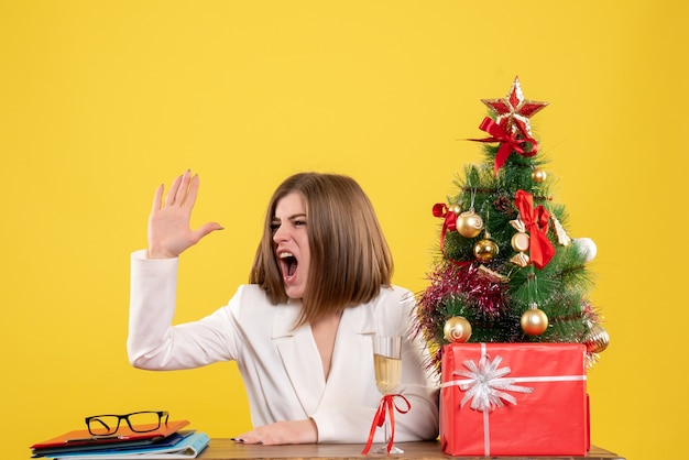 Ärztin der Vorderansicht, die vor Tisch auf gelbem Hintergrund mit Weihnachtsbaum und Geschenkboxen sitzt