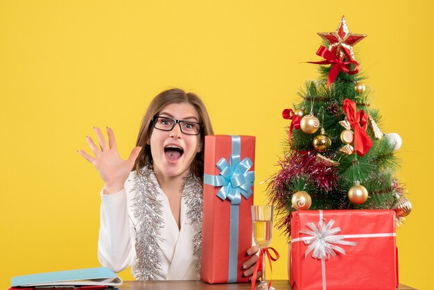 Ärztin der Vorderansicht, die vor Tisch mit Geschenken und Baum auf gelbem Hintergrund mit Weihnachtsbaum und Geschenkboxen sitzt