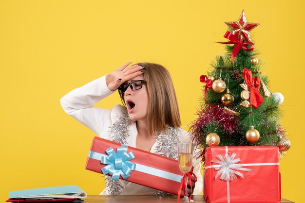 Ärztin der Vorderansicht, die vor Tisch mit Geschenken und Baum auf gelbem Hintergrund mit Weihnachtsbaum und Geschenkboxen sitzt