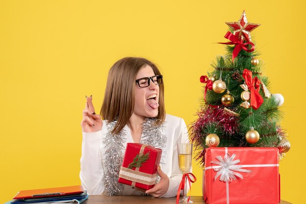 Ärztin der Vorderansicht, die vor Tisch mit Geschenken und Baum auf gelbem Hintergrund mit Weihnachtsbaum und Geschenkboxen sitzt