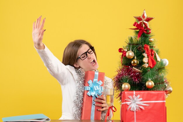 Ärztin der Vorderansicht, die vor Tisch mit Geschenken und Baum auf gelbem Hintergrund mit Weihnachtsbaum und Geschenkboxen sitzt