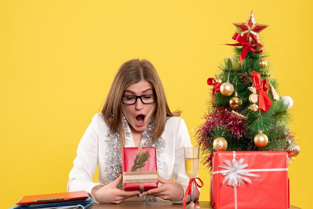 Ärztin der Vorderansicht, die vor Tisch mit Geschenken und Baum auf gelbem Hintergrund mit Weihnachtsbaum und Geschenkboxen sitzt