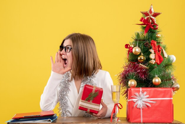 Ärztin der Vorderansicht, die vor Tisch mit Geschenken und Baum auf gelbem Hintergrund mit Weihnachtsbaum und Geschenkboxen sitzt