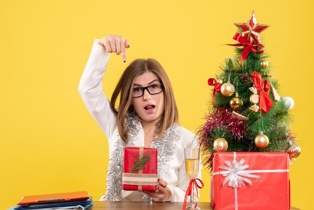 Ärztin der Vorderansicht, die vor Tisch mit Geschenken und Baum auf gelbem Hintergrund mit Weihnachtsbaum und Geschenkboxen sitzt