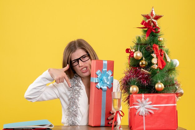 Ärztin der Vorderansicht, die vor Tisch mit Geschenken und Baum auf gelbem Hintergrund sitzt