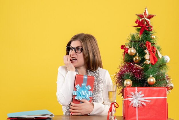 Ärztin der Vorderansicht, die vor Tisch mit Geschenken und Baum auf gelbem Hintergrund sitzt