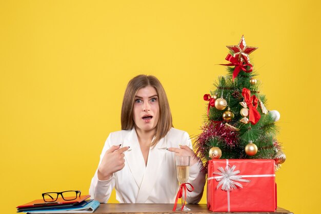 Ärztin der Vorderansicht, die vor Tisch mit Weihnachtsgeschenken und Baum auf gelbem Hintergrund sitzt