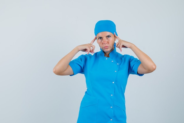 Ärztin, die Finger auf Schläfen in blauer Uniform hält und nachdenklich, Vorderansicht schaut.