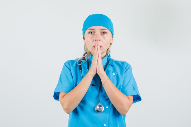 Ärztin, die Hände in der Gebetsgeste in der blauen Uniform hält und ängstlich aussieht.