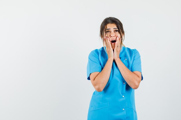 Ärztin, die Hände in der Nähe von offenem Mund in blauer Uniform hält und überrascht aussieht