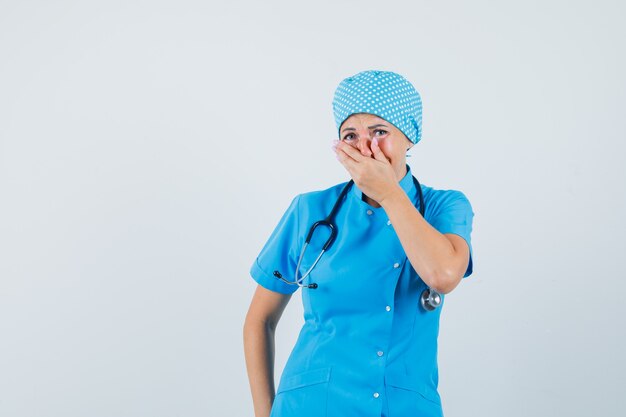 Ärztin, die Hand auf Mund in blauer Uniform hält und froh aussieht. Vorderansicht.