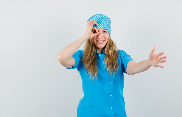 Ärztin, die Hand mit ok Zeichen auf Auge in blauer Uniform streckt und fröhlich schaut.
