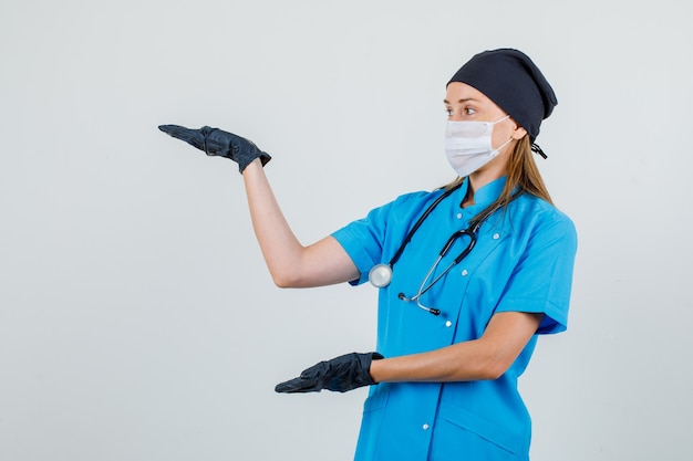 Ärztin, die Handflächen offen hält, um etwas in Uniform, Handschuhen, Maskenvorderansicht zu zeigen.