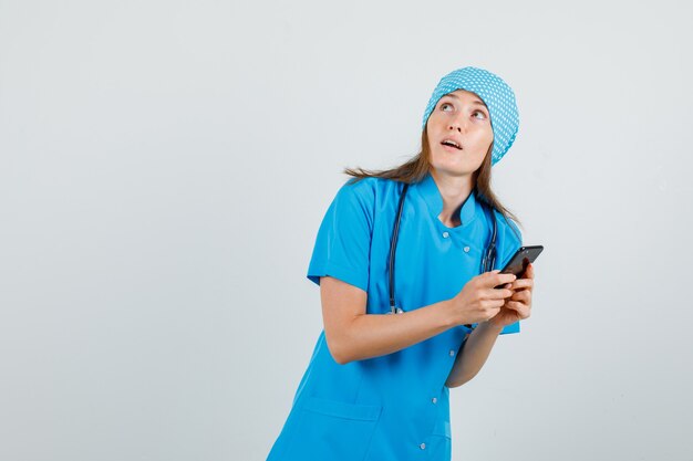 Ärztin, die nach oben schaut, während Smartphone in blauer Uniform hält und konzentriert schaut