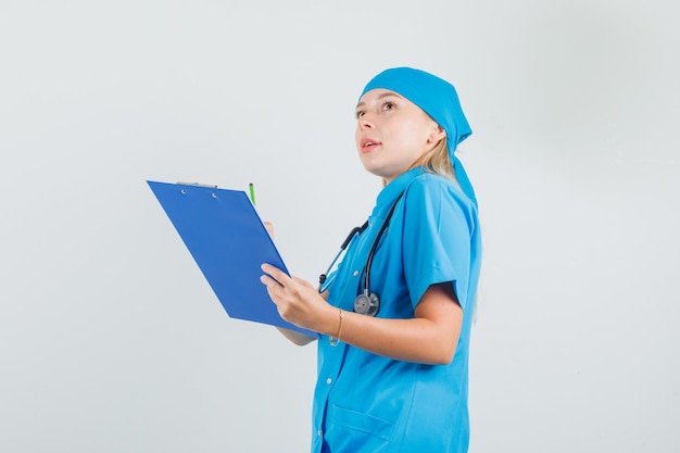 Ärztin, die nach oben schaut, während Zwischenablage und Bleistift in der blauen Uniform halten