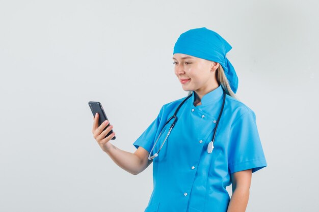 Ärztin, die Smartphone in blauer Uniform hält und betrachtet und fröhlich schaut.