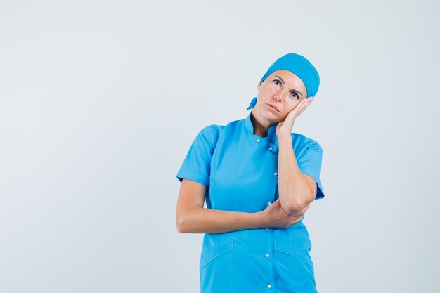 Ärztin, die Wange auf Handfläche in blauer Uniform lehnt und nachdenklich aussieht. Vorderansicht.