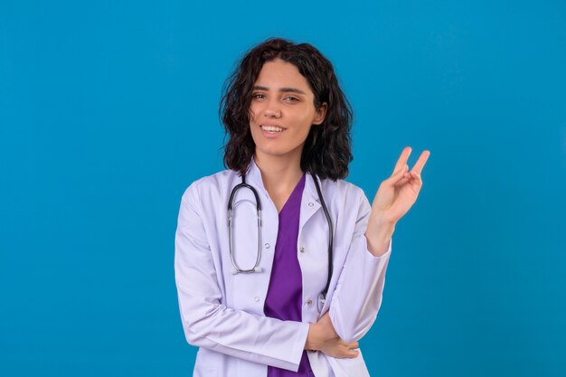 Ärztin, die weißen Kittel mit dem Stethoskop trägt, das fröhlich lächelt, beiseite schaut und Nummer zwei oder Siegeszeichen mit den Fingern auf isoliertem Blau zeigt