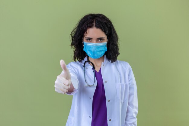 Ärztin, die weißen Kittel mit Stethoskop in der medizinischen Schutzmaske mit Lächeln auf Gesicht trägt, zeigt Daumen oben stehend auf lokalisiertem Grün