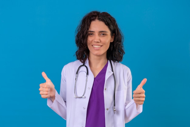 Ärztin, die weißen Kittel mit Stethoskop mit glücklichem Lächeln trägt, zeigt Daumen hoch stehend auf isoliertem Blau