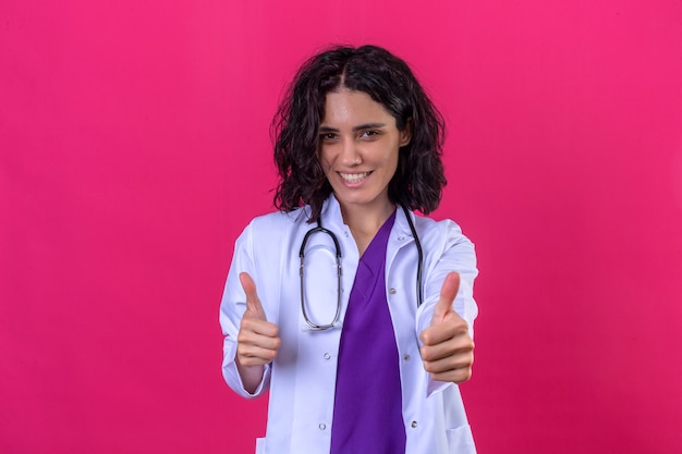 Ärztin, die weißen Kittel mit Stethoskop mit glücklichem Lächeln trägt, zeigt Daumen hoch stehend auf lokalisiertem Rosa