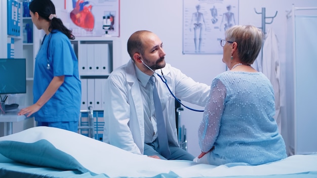 Ärztin für ältere Frauen in einer modernen Privatklinik, während sie auf dem Krankenhausbett sitzt und der Arzt ihren Herzschlag hört. Heilmedizinische Behandlung, Facharztberatung