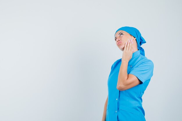 Ärztin in blauer Uniform berührt ihre Haut auf der Wange und sieht nachdenklich aus, Vorderansicht.