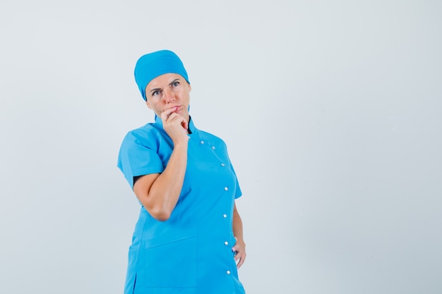 Ärztin in blauer Uniform, die Hand am Kinn hält und nachdenklich, Vorderansicht schaut.