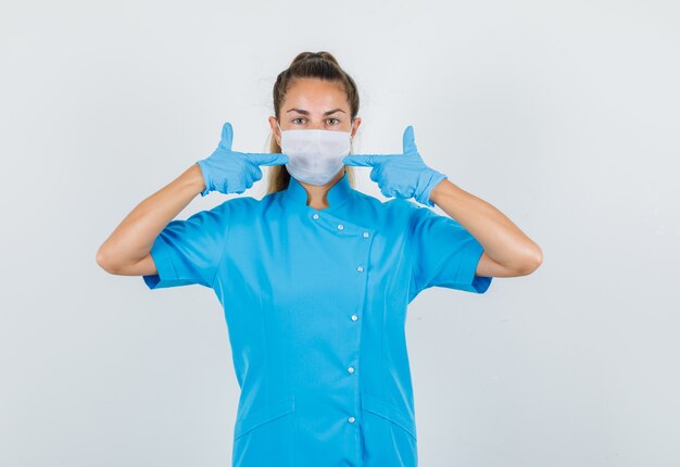 Ärztin in blauer Uniform, Handschuhe zeigen auf ihre Maske und sehen vorsichtig aus