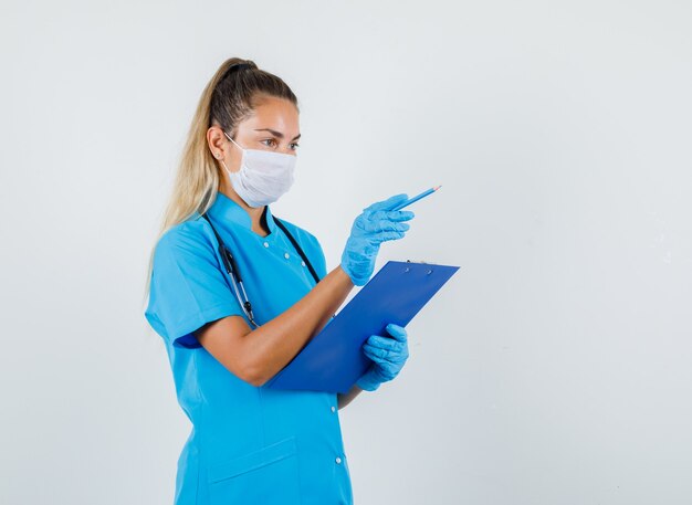 Ärztin in blauer Uniform, Maske, Handschuhe zeigen Bleistift weg, während sie Zwischenablage hält