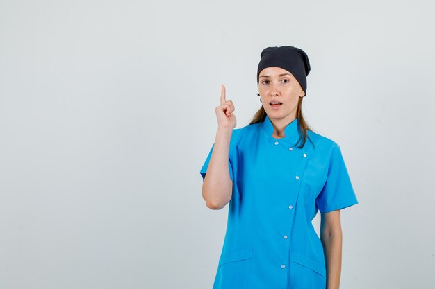Ärztin in blauer Uniform, schwarzer Hut zeigt mit dem Finger nach oben und sieht selbstbewusst aus