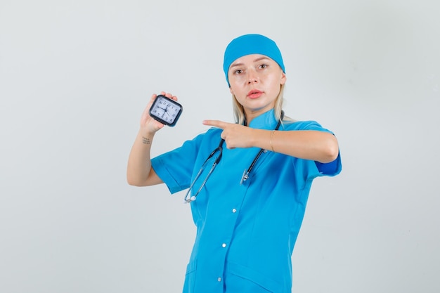 Ärztin in blauer Uniform zeigt auf die Uhr und sieht ernst aus