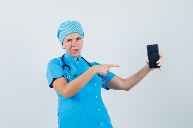 Ärztin in blauer Uniform zeigt auf Handy und schaut verwirrt, Vorderansicht.